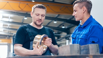 <br />
Zwei Personen in Overalls im Werkshalle stehend im Gespräch miteinander: Eine Person hält breites metallenes Verbundstück in Händen und blickt darauf sprechend