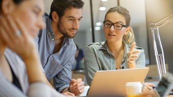 Fokus auf zwei Personen, die sich unterhalten und dabei auf einen Laptopbildschirm blicken, im Vordergrund sitzt eine weitere Person in der Unschärfe