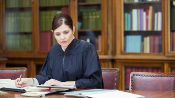 Person in schwarzer Richterrobe sitzt an einem Tisch über Unterlagen und macht Notizen, im Hintergrund verschwommen Bücherregale.