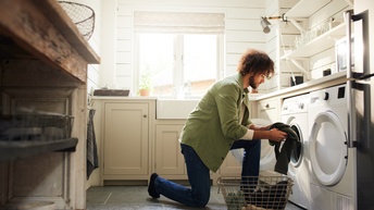 Bärtige Person mit Locken kniet mit einem Bein am Boden, das andere angewinkelt aufgestellt, vor Waschmaschine und hält Wäsche in Händen, daneben Wäschetrockner, vor Person Drahtkorb mit Wäsche, ringsum Waschküchenregale und Fenster