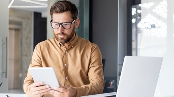 Eine Person mit Brille, kurzen, braunen Haaren und braunem Bart sitzt an einem weißen Tisch. In den Händen hält sie ein Tablet, auf das sie blickt. Links vor der Person ist ein aufgeklappter Laptop
