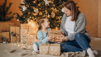 Erwachsene Person übergibt einem Kind ein Geschenk, im Hintergrund steht ein geschmückter Weihnachtsbaum und weitere Geschenke sind daneben platziert