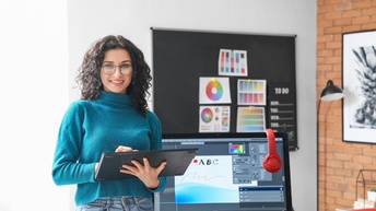 Porträt einer Person mit lockigen, dunklen Haare und Brille, die ein Tablet hält. Hinter der Person sind ein Bildschirm und ein aufgeklappter Laptop. Auf dem Bildschirm ist ein Programm geöffnet zur Bearbeitung von Dateien