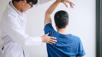 Person in weißer Arbeitskleidung mit Brille führt bei einer anderen Person therapeutische Untersuchung der Hand und Schulter einer anderen Person durch