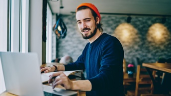 Person mit dunklen Haaren, Bart und oranger Mütze steht an einer Theke bei einem Fenster in einem Lokal und blickt auf einen geöffneten Laptopbildschirm