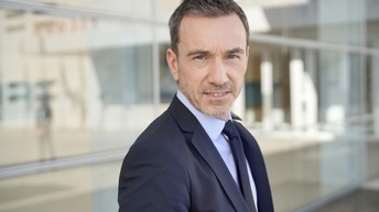 Halbkörperportrait einer Person in Anzug, im Hintergrund spiegelnde Gebäudefassade