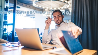 Person lehnt in Schreibtischstuhl, hält in einer Hand Stift in Nähe des Kopfes und blättert mit anderer Hand durch Dokumentenmappe, vor ihr aufgeklapptes Notebook platziert