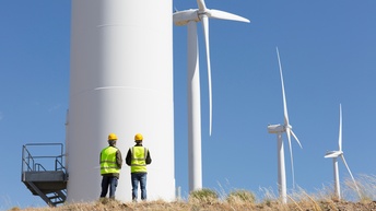 Zwei Personen mit gelben Helmen in Warnwesten stehen vor Windrad und blicken auf weitere, dahinter nachfolgende Windräder unter blauem Himmel