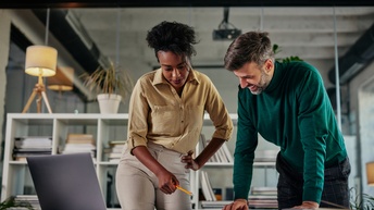 Zwei Personen stehen bei einem Schreibtisch und blicken auf Unterlagen, dahinter zeigt sich ein modernen Büro mit einem Regal und Unterlagen