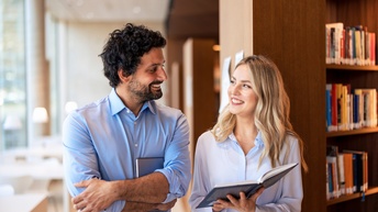 Zwei Personen in Bibliothek stehend lächeln einander an, eine Person hält aufgeklapptes Buch in Händen