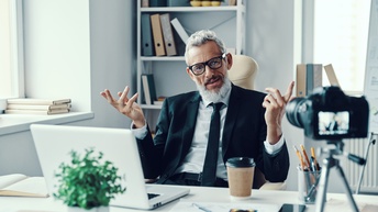 Auf einem Tisch steht eine Halterung, in der horizontal ein Handy fixiert ist. Das Handy filmt eine Person, die hinter einem Schreibtisch sitzt, in die Kamera spricht und mit den Händen gestikuliert. Vor der Person ist ein Laptop