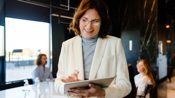 Porträt lächelnden Person mit Brillen, die auf Tablet tippt und blickt, im Hintergrund verschwommen Besprechungsraum mit weiteren Personen
