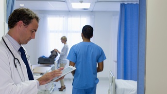 Seitliche Aufnahme einer Person in einem weißen Kittel, mit Stethoskop und mit Tablet in der Hand, auf das sie blickt. Im Hintergrund geht eine Person in blauer Kleidung sowie packt eine andere Person einen Koffer aus