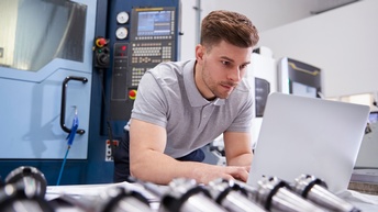 Person arbeitet mit einem Laptop in einer Werkstätte, im Hintergrund befinden sich verschiedene Maschinen 