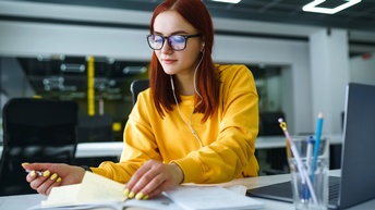 Person mit Brillen und gelbem Pullover sitzt an Tisch mit Kopfhörern in den Ohren und durchblättert Unterlagen, aufgeklapptes Notebook und Glas mit Stiften nebenstehend