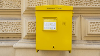 Ein gelber Kasten hängt an einer Hausfassade. Auf dem Kasten steht Post