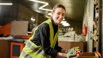 Lächelnde Person in gelber Warnweste mit Handschuhen beugt sich über Karton und hievt Flaschenverschlüsse aus Plastik heraus