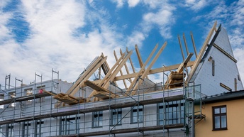 Vor einem mehrstöckigen Gebäude steht ein Gerüst. Die Fassade besteht aus Dämmmaterial. Der Dachboden besteht aus schrägen Holzbalken sowie ein paar stehenden Ziegelmauern. Auf dem Boden des Ausbaus liegen Holzbretter