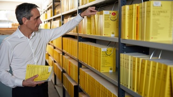 Person mit weißem Hemd hält ein Rechtsbuch in Händen und greift mit der anderen Hand zu einem weiteren Buch in einem Regal einer Bibliothek