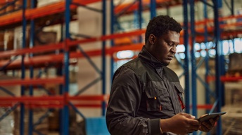 Seitenansicht einer Person in Arbeitskleidung in einer Lagerhalle. Die Person hält ein Tablet in den Händen und wirkt konzentriert. Im Hintergrund sind hohe Regale zu sehen.