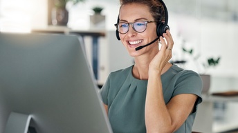 Person mit kurzen dunklen Haaren und Brille blickt auf einen Computerbildschirm. Auf dem Kopf hat sie ein schwarzes Headset mit Mikrofon