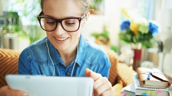 Eine Person mit Brille und Kopfhörern sitzt auf einem Sofa und hält ein Tablet, auf das sie blickt. Die Person trägt eine Brille und Kopfhörer. Neben ihr liegen Bücher
