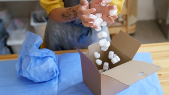 Detailansicht der Hände einer Person, die Füllmaterial in einen Verpackungskarton fallen lässt.