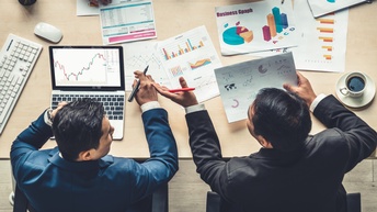 Zwei Personen in Businesskleidung werden aus der Vogelperspektive sitzend an einem Tisch gezeigt wie sie Statistiken am Papier und am Laptop besprechen, die Hände einer dritten Person ragen von der anderen Seite des Tisches herein