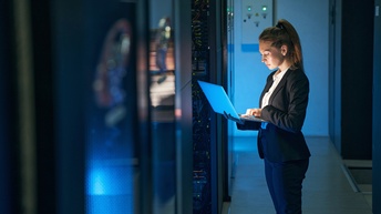Person in Businesskleidung steht in einem Serverraum und hält einen Laptop in der Hand