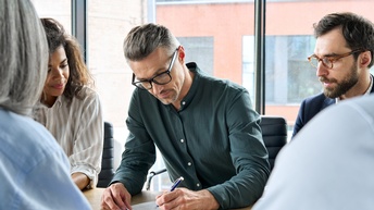Mehrere Personen sitzen um Besprechungstisch und blicken auf eine Person, die Dokument unterzeichnet