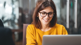 Portrait von einer lächelnden Person mit dunklen schulterlangen Haaren und Brille sowie gelbem Pullover, die vor einem Laptop sitzt
