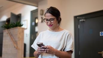 Person mit Brille steht mit einem Smartphone in einem Wohnraum und blickt fröhlich darauf