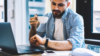 Eine Person mit kurzen Haaren und Bart sitzt an einem Tisch und blickt auf ihre Armbanduhr. Vor ihr steht ein aufgeklappter Laptop. In der rechten Hand hält sie eine Tasse. Neben dem Laptop ist ein aufgeklapptes Notizbuch.