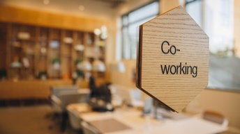 Holzschild mit Aufschrift Co-Working auf einer Glasfront montiert, im Hintergrund zeigt sich eine beleuchtete Büroräumlichkeit in der Unschärfe