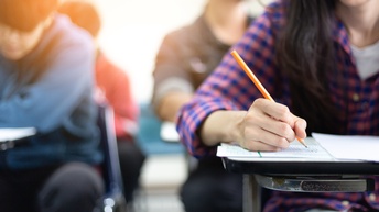 Personen sitzen an Schreibtischen und schreiben während des Unterrichts auf Zetteln, Fokus auf eine Person, die Stift in Hand hält.