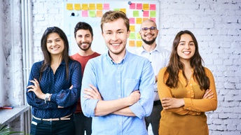 Porträt von fünf versetzt neben- und hintereinander stehenden Personen, die vor einer Ziegelmauer stehen. Hinter den Personen hängt an der Wand eine Tafel mit Zetteln