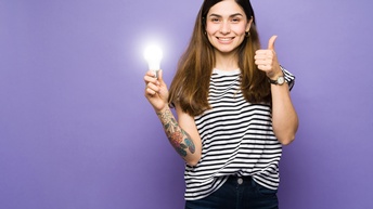 Porträt einer lächelnden Person mit offenen, dunklen Haaren, Jeans, gestreiftem Oberteil sowie Ohrringen, die vor einer lila Wand steht und den linken Daumen nach oben hält. In der rechten Hand hält die Person eine leuchtende Glühbirne