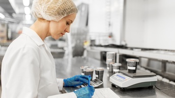 Person mit weißem Haarnetz, weißer Arbeitsbekleidung und blauen Handschuhen kontrolliert eine Liste und tippt in einem Taschenrechner ein, während Becher auf einer Waage stehen