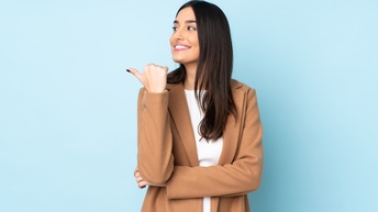 Porträt von einer lächelnden Person mit langen dunklen Haaren, die mit der Hand nach links zeigt und blickt und vor einem hellblauem Hintergrund steht
