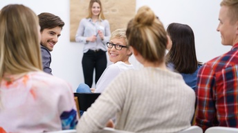 Rückenansicht, sechs neben- und hintereinander sitzender Personen, die teilweise nach vorne blicken und sich gegenseitig anblicken. Ihnen gegenüber steht eine weitere Person vor einer Wand, auf der ein großes, braunes Stück Papier hängt