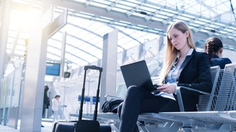 Person sitzt auf Sitzgelegenheiten in Bahnterminal und hat aufgeklapptes Notebook am Schoß, nebenstehend Rollkoffer