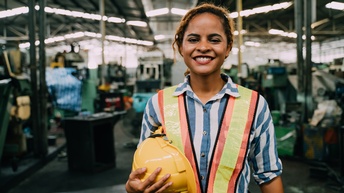 Halbporträt einer Person mit Sicherheitsweste und Helm in der rechten Hand. Im Hintergrund ist eine hohe Halle mit Geräten