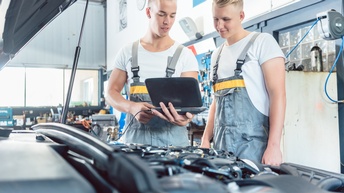 Zwei Personen in Arbeitslatzhosen stehen in Werkstatt vor Auto mit geöffneter Motorhaube und blicken auf Computer