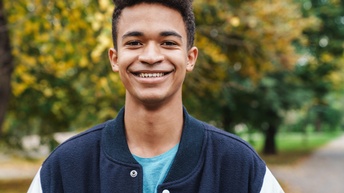 Halbporträt einer Person mit Übergangsjacke und kurzen, dunklen, gelockten Haaren. Im Hintergrund sind ein asphaltierter Weg und leicht verfärbte Bäume