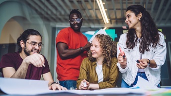 Zwei Personen sitzen, zwei Personen stehen an einem Tisch. Vor ihnen ist ein großes Plakat. Eine Person blickt darauf. Eine Person hält einen Zettel. Zwei weitere Personen halten einen Stift