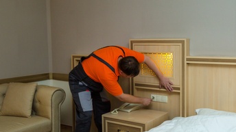 Eine Person in Latzhose steht neben einer Wand, beugt sich nach vorne und hält die rechte Hand vor eine weiße Steckdose, die über einem Kästchen ist. Das Kästchen steht neben einem Bett.