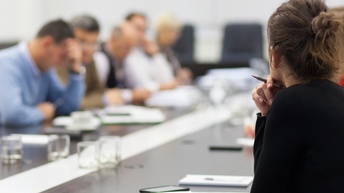 Rückenansicht einer Person mit zum Dutt gebundenen Haaren, Hand mit Kugelschreiber an Kinn gestützt, im Hintergrund verschwommen weitere Personen an Tisch sitzend