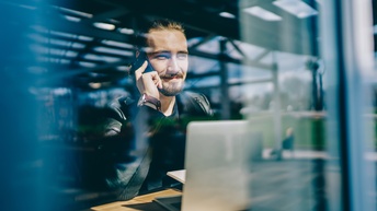 Junger Mann am Smartphone arbeitet mit Laptop in einem Restaurant vor einem Fenster