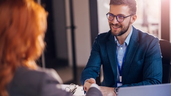 Person mit kurzen dunklen Haaren, Bart und Brille sowie Anzug sitzt an einem Tisch mit Laptop und Unterlagen und gibt einer Person, die gegenüber in der Unschärfe sitzt freudig die Hand