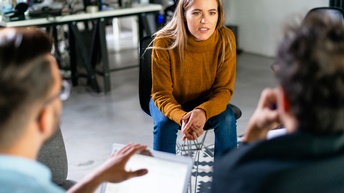 Person mit braunem Pullover und langen blonden Haaren sitzt auf einem Sessel und spricht zu anderen Personen, die ihr in der Unschärfe gegenübersitzen